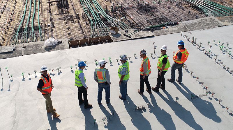 A row of construction managers stand looking off towards their construction site at mid-day.